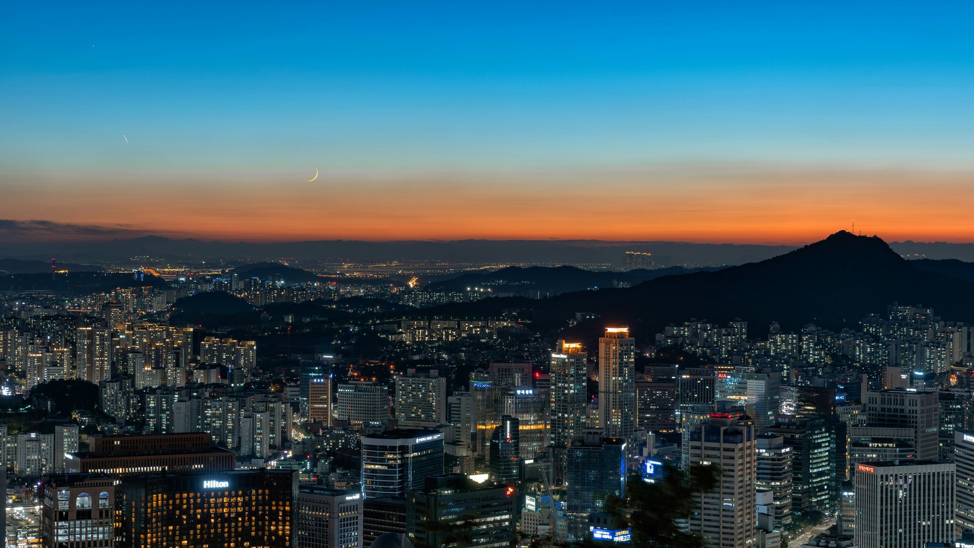 Seoul skyline at dusk beneath a crescent moon